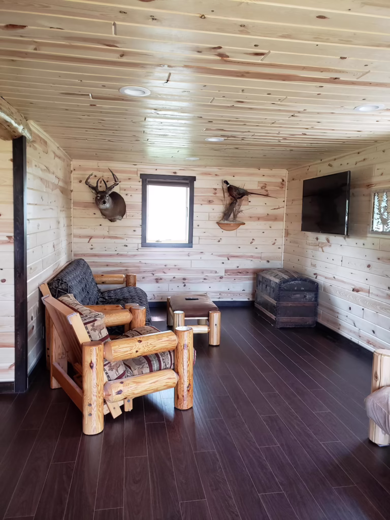 Rustic living room with wood-paneled walls and ceiling, log furniture, mounted deer head, TV on the wall, wood floor, and a small trunk as a coffee table. Natural light from a central window brings in the peaceful ambiance of a cozy Rainy Lake outpost—perfect for relaxing after a long day of fishing. | Rainy Lake, MN